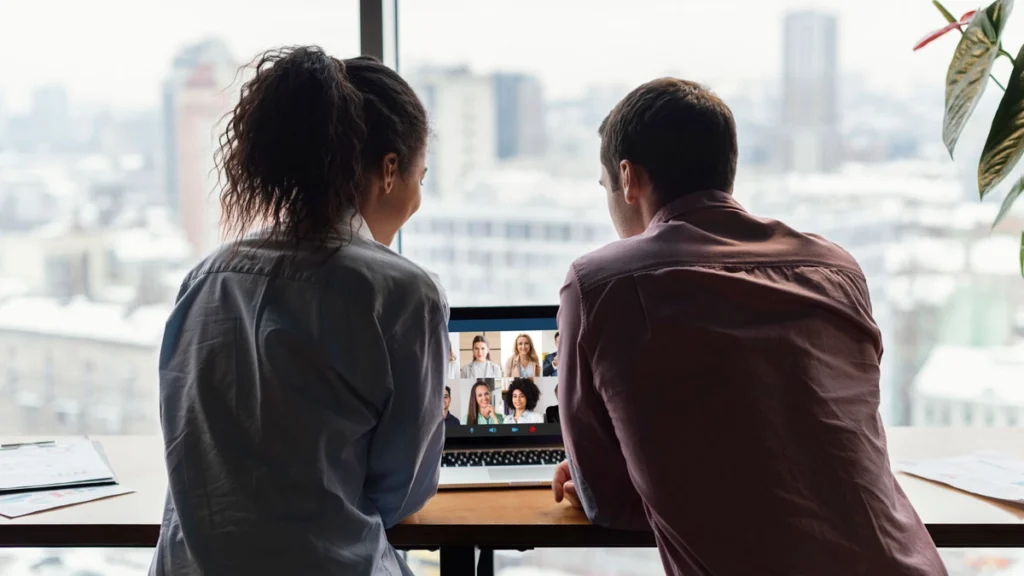 Two people sitting at a table with a laptop on a video call, overlooking a cityscape in the background.