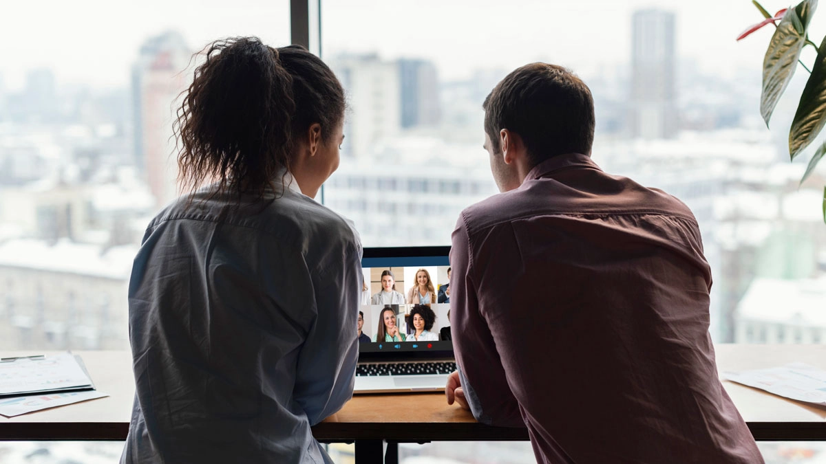 Two people sitting at a table with a laptop on a video call, overlooking a cityscape in the background.