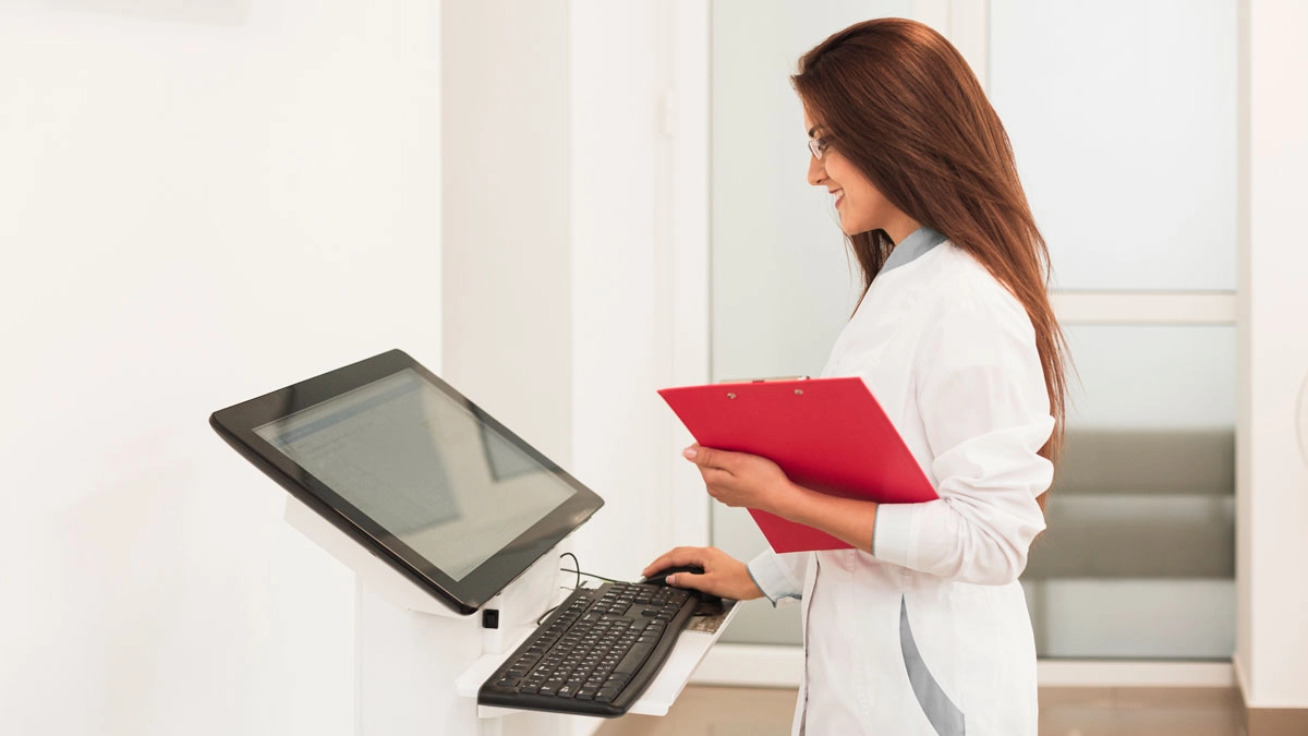 A woman in a white lab coat stands beside a computer, focused on her work in a laboratory setting.