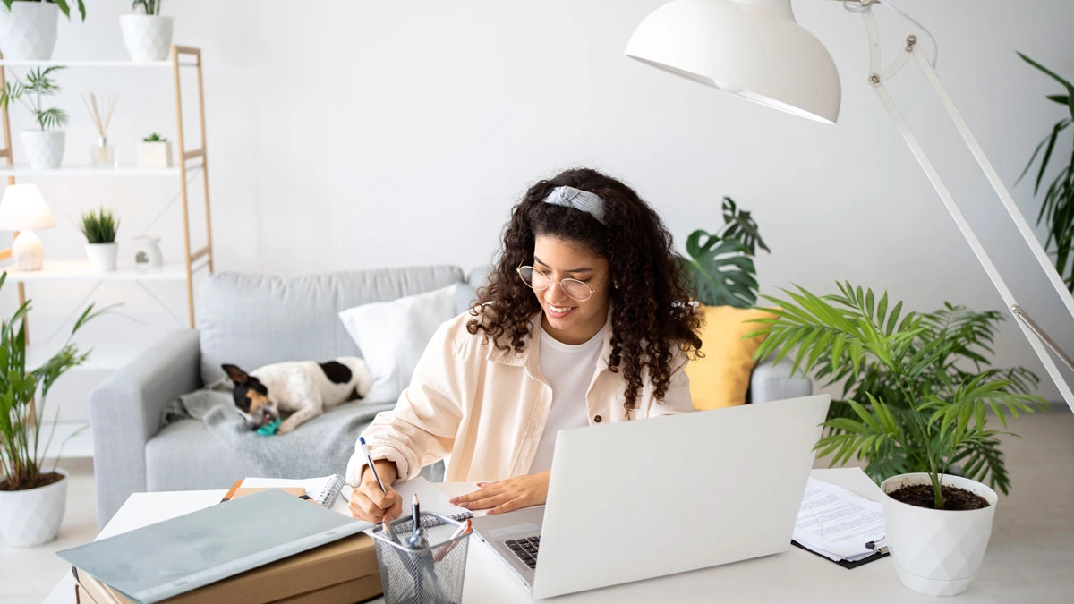 A woman sits at a desk with a laptop and a dog in a cozy home office setting.